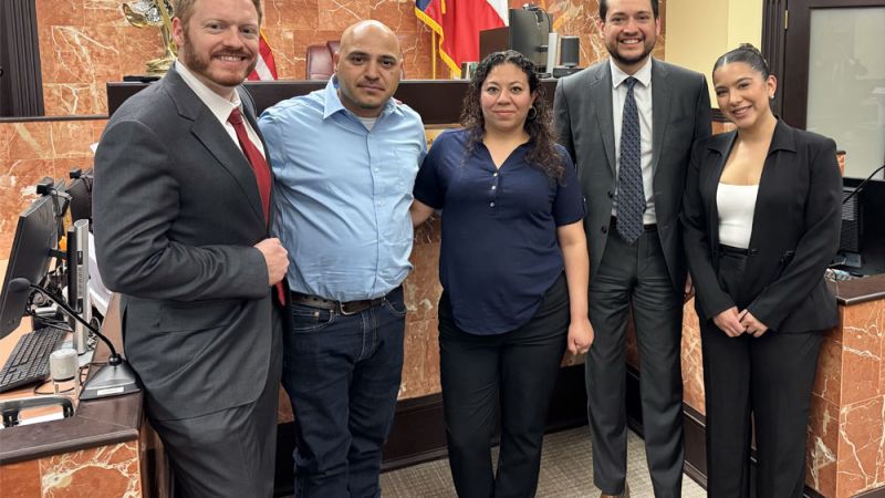 Group of professionals smiling in a courtroom setting, with flags and a seal in the background.