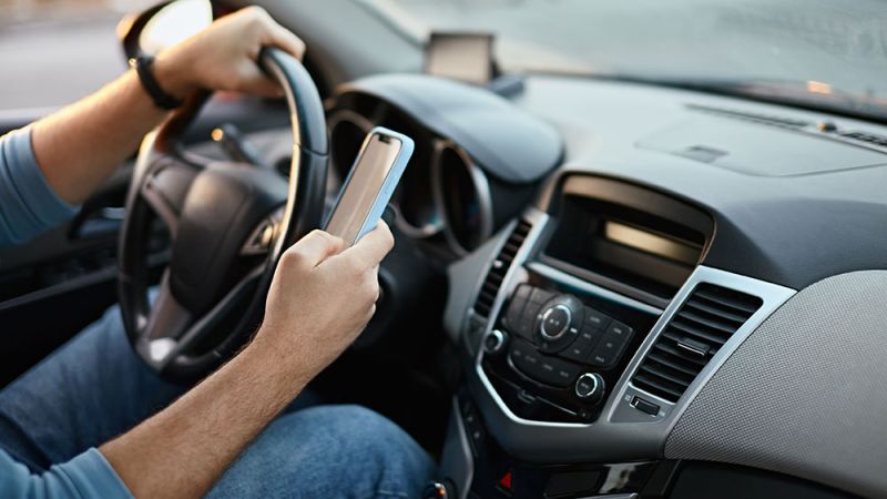 Driver holding a smartphone while steering a car, illustrating distracted driving hazards.