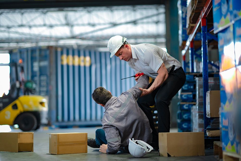 Warehouse safety: worker assists another after a fall among boxes and forklift. Safety helmets nearby.