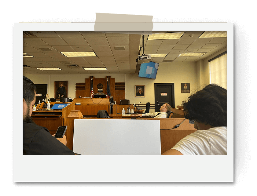 Courtroom scene with people on phones and one person leaning back, empty seats and a judge in the background.