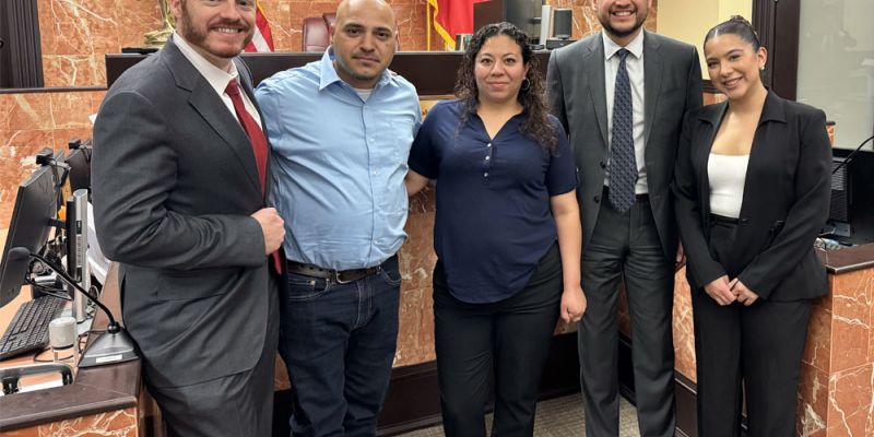 Group of professionals smiling in a courtroom setting, with flags and a seal in the background.