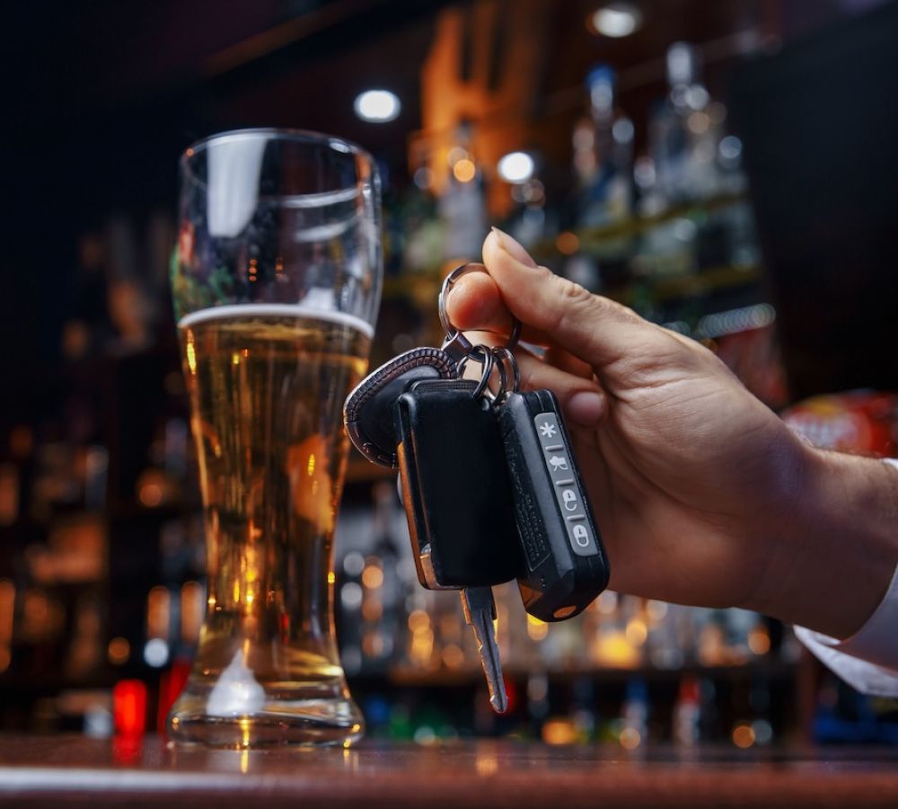 A hand holds car keys beside a tall glass of beer, situated on a bar counter surrounded by blurred bottles and dim lighting.