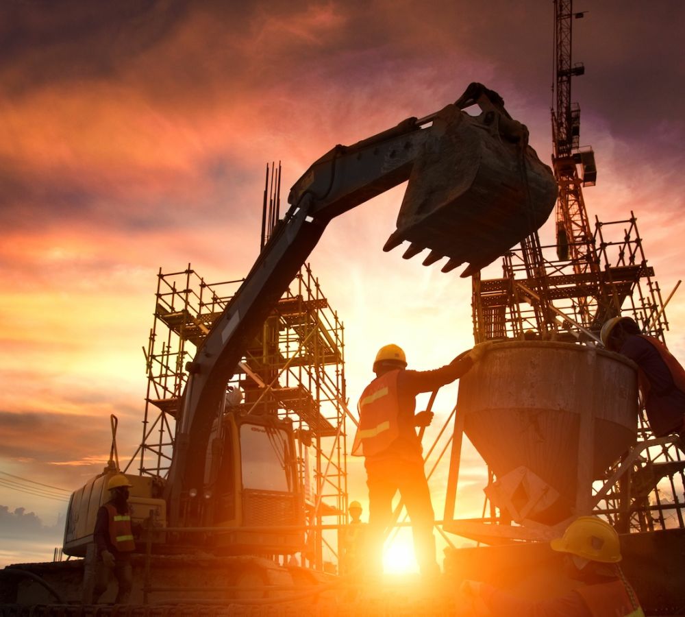 Construction workers and machinery on a building site at sunset, silhouetted against a colorful sky.