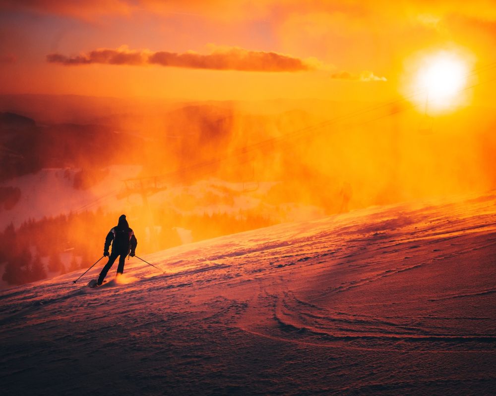 Skier descends snowy mountain at sunset, vibrant orange glow illuminating the scene.