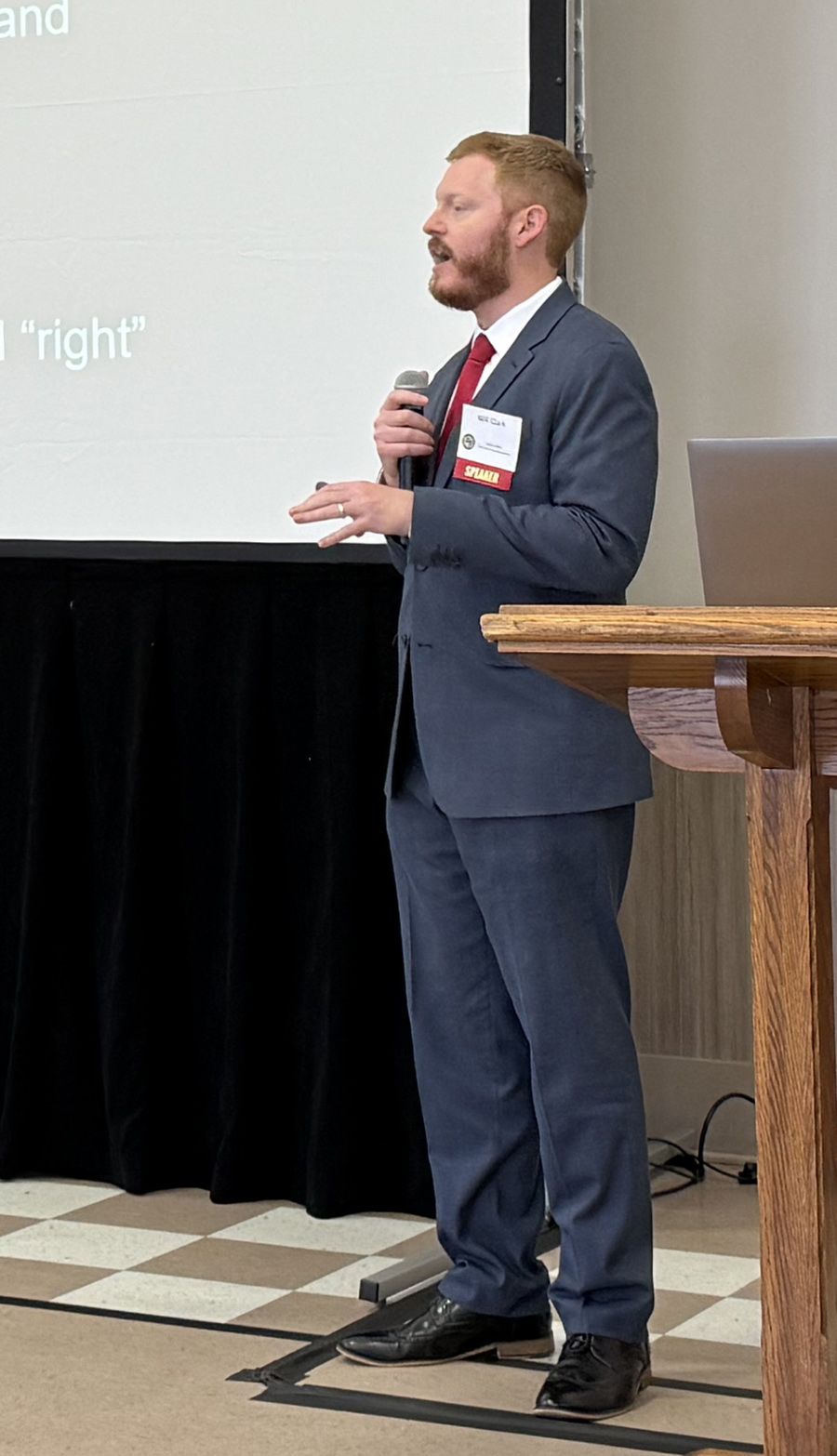 Man in a suit delivers a presentation with a microphone, standing by a podium and a projector screen.