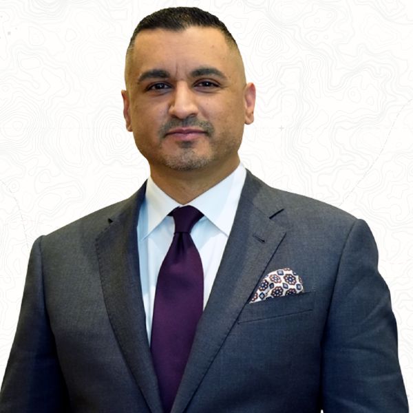 Business professional in a suit and tie, standing against a white background with a subtle pattern.