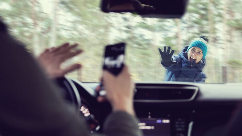 Distracted driver using phone, startled woman outside windshield, winter forest setting.