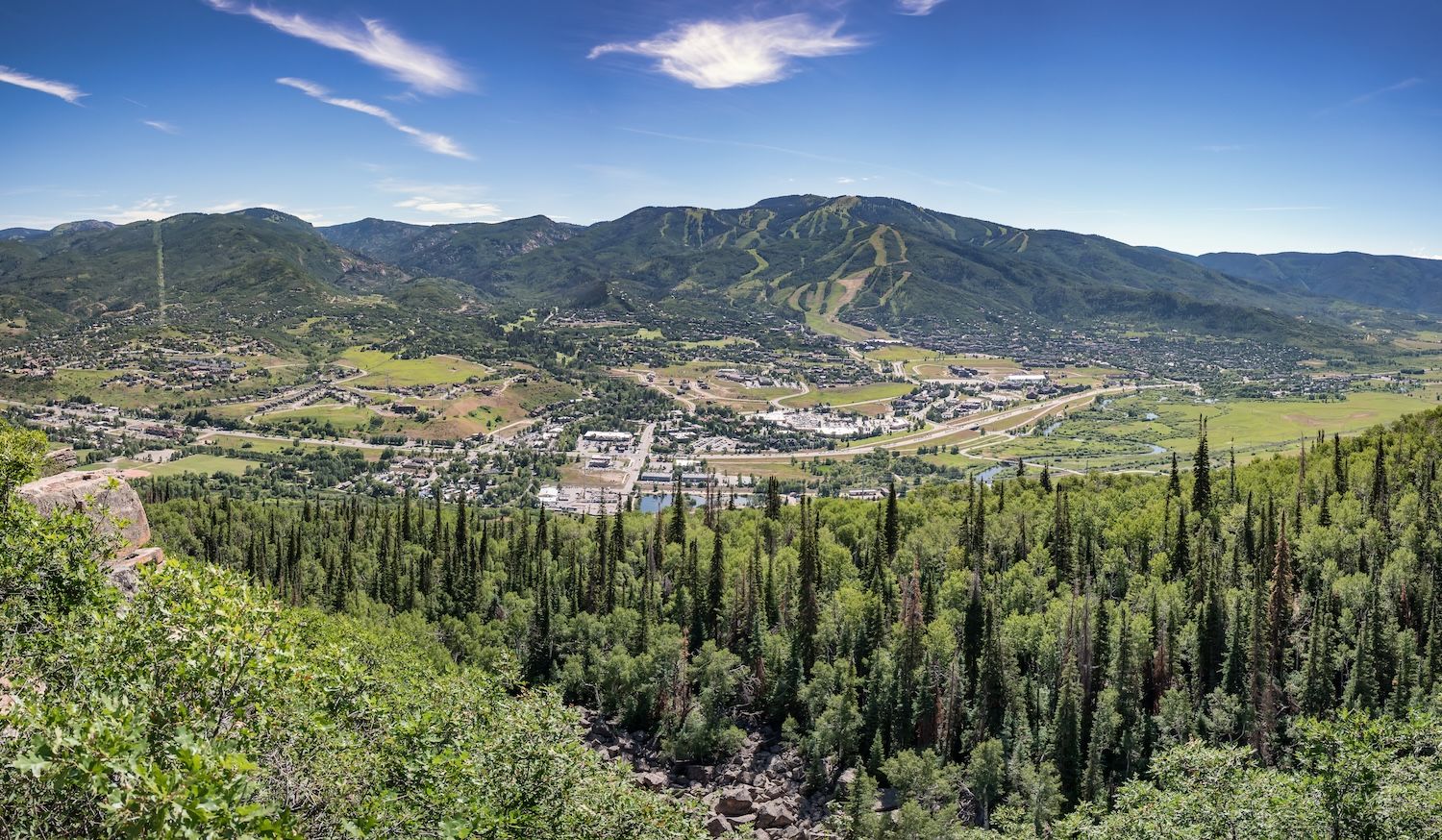 Panoramic view of mountain landscape with lush green forests and a quaint town nestled in a sunny valley below.