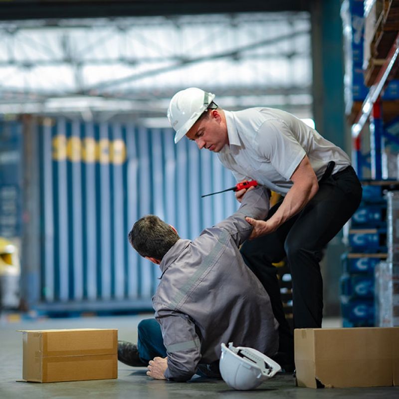 Warehouse safety: worker assists another after a fall among boxes and forklift. Safety helmets nearby.