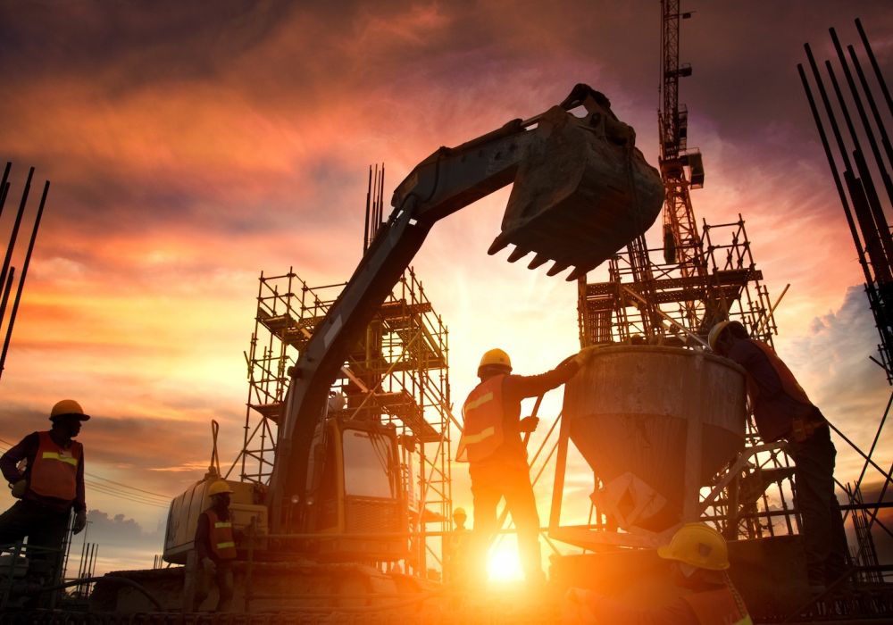 Construction workers and machinery on a building site at sunset, silhouetted against a colorful sky.