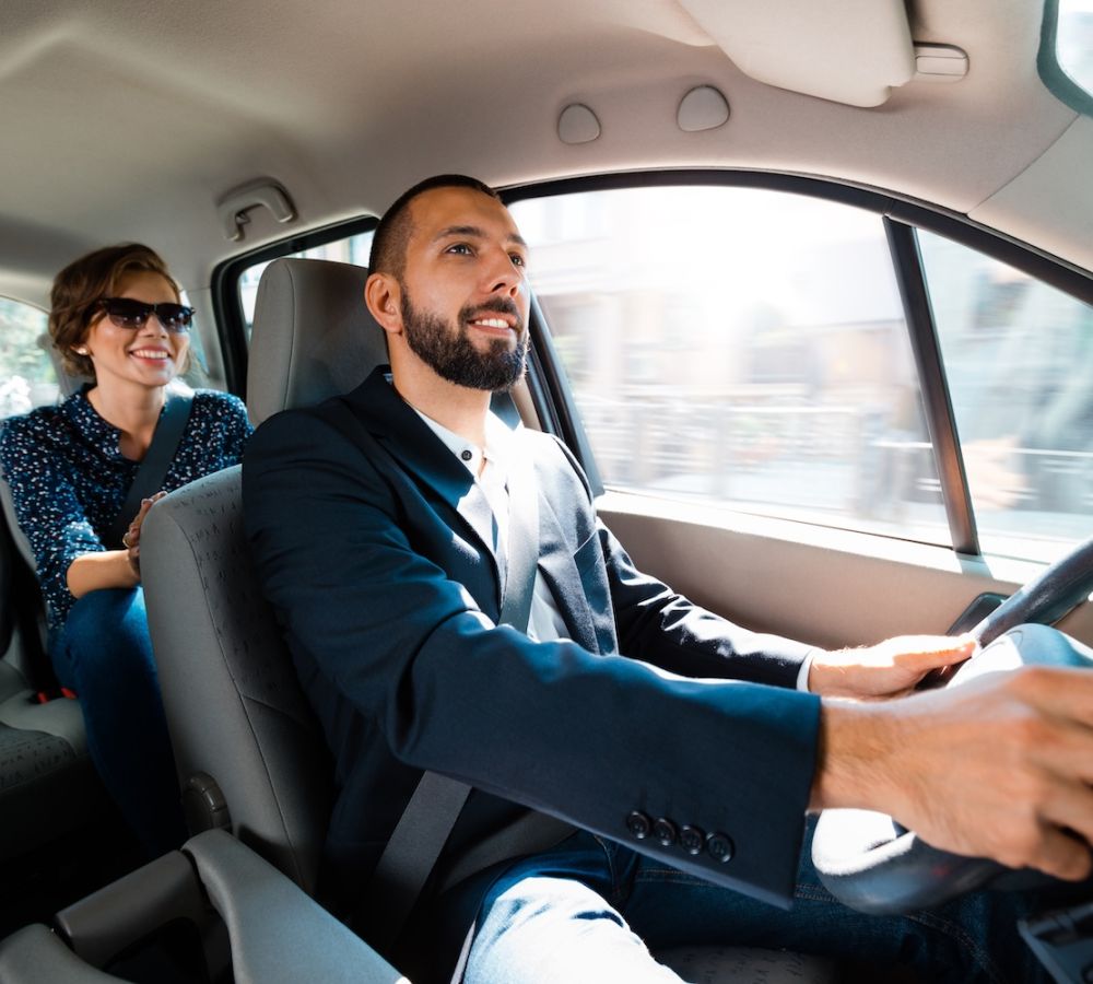Man driving car with a smiling passenger in the back seat, showcasing a pleasant ride-sharing experience.