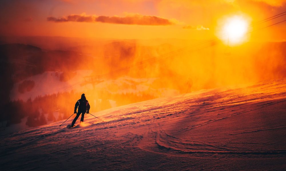 Skier descends snowy mountain at sunset, vibrant orange glow illuminating the scene.