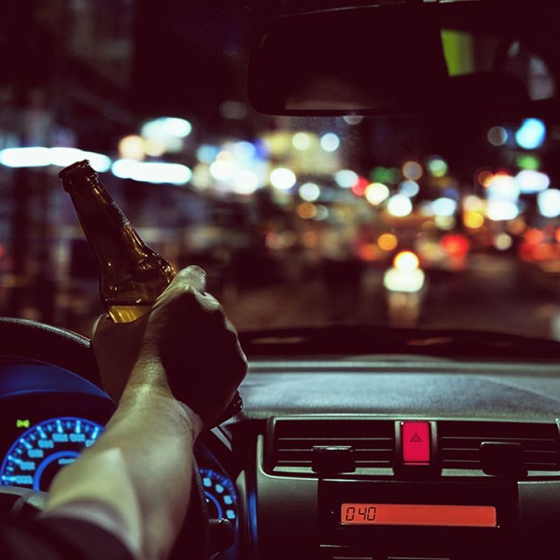 Driver holds beer bottle in car at night, emphasizing dangers of drunk driving and road safety concerns.