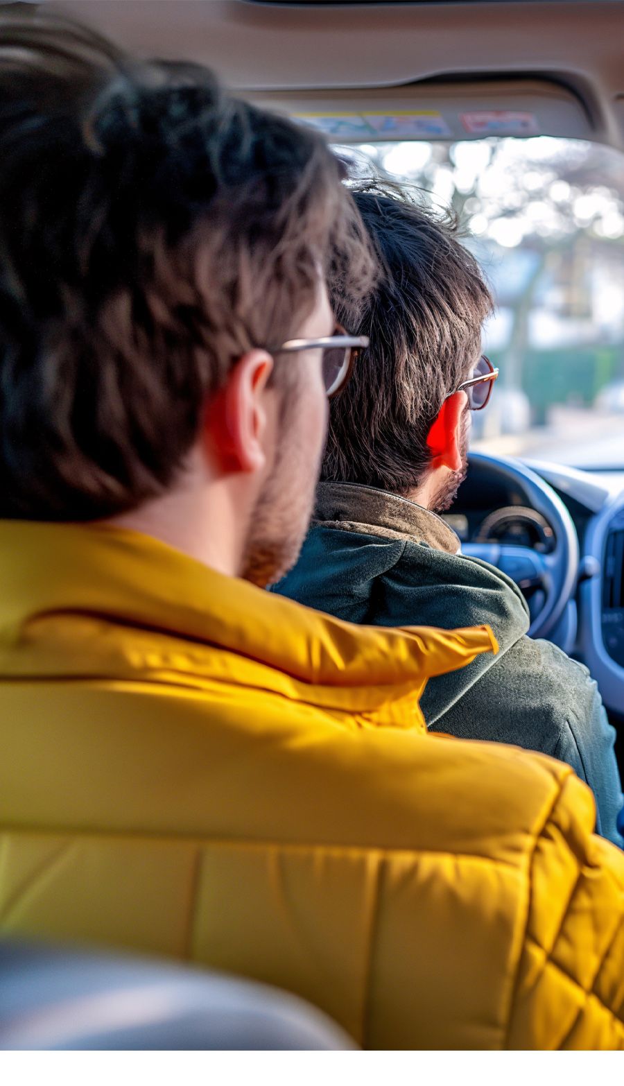 Two people wearing sunglasses in a car, with one driving, captured from the backseat perspective.