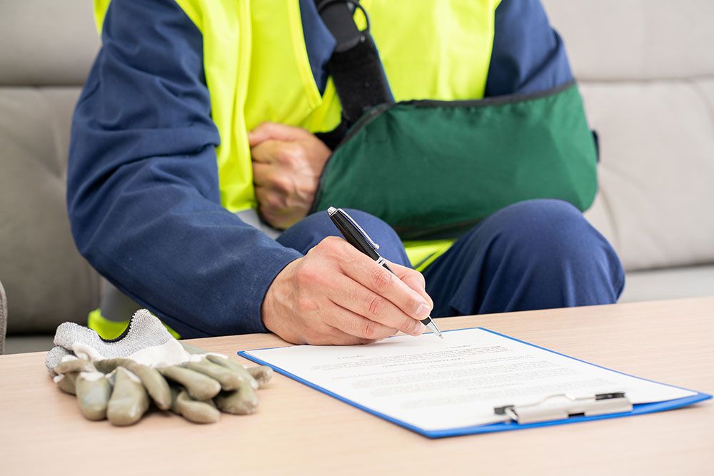 Injured worker with arm sling signing documents on clipboard, work gloves nearby.