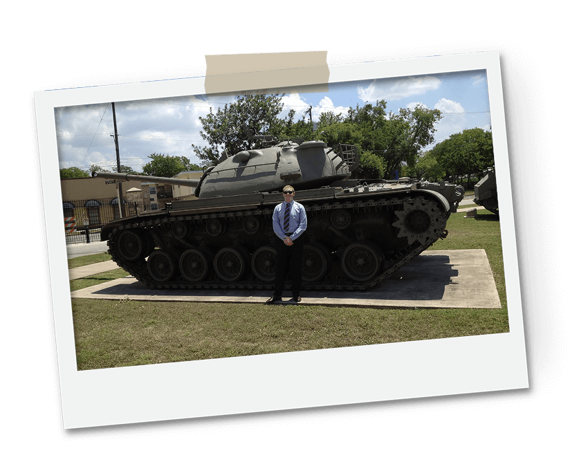 Man in formal attire standing in front of a large tank on display outside, under a clear blue sky.