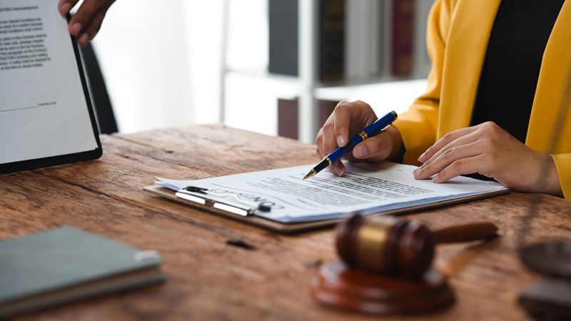 Person in yellow jacket signing document at desk with gavel and tablet, legal paperwork concept.