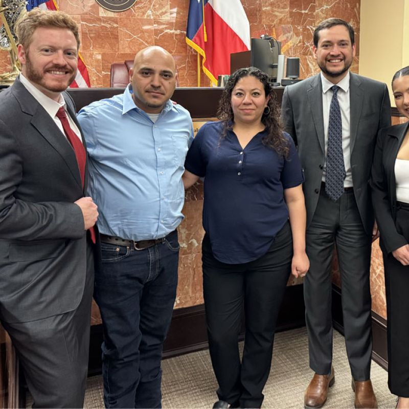 Group of professionals smiling in a courtroom setting, with flags and a seal in the background.
