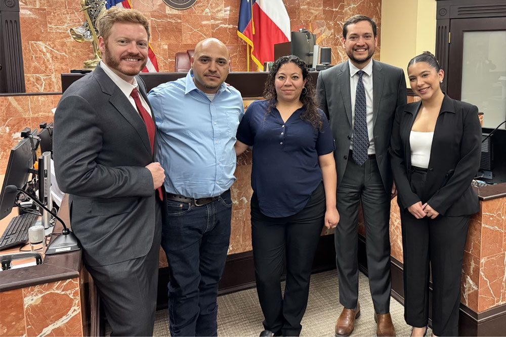 Group of professionals smiling in a courtroom setting, with flags and a seal in the background.