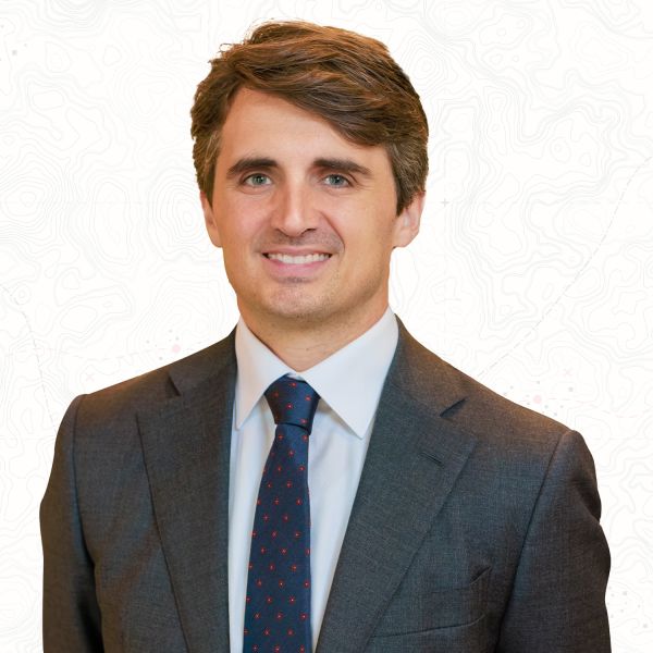 Smiling man in a suit and tie, standing against a white, patterned background.