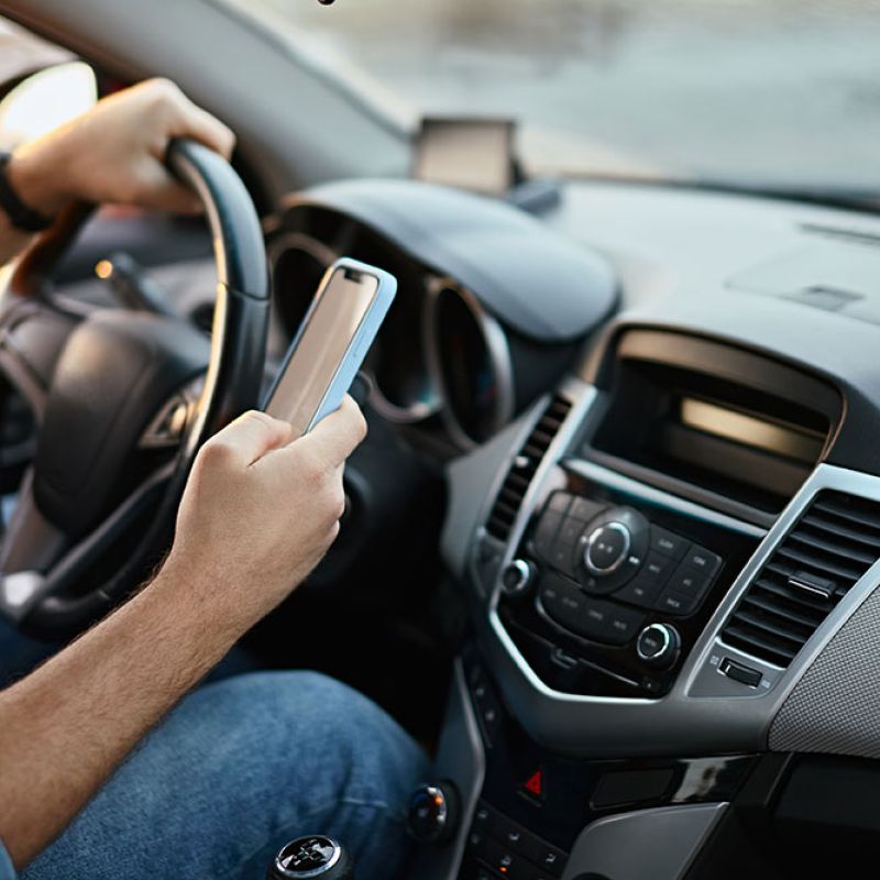 Driver holding a smartphone while steering a car, illustrating distracted driving hazards.