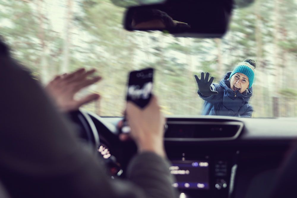 Distracted driver using phone, startled woman outside windshield, winter forest setting.