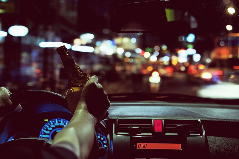 Driver holds beer bottle in car at night, emphasizing dangers of drunk driving and road safety concerns.