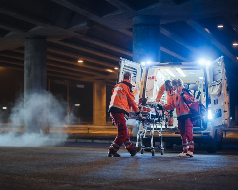 Emergency medical team loading a patient into an ambulance under a bridge at night.