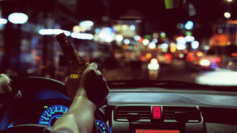 Driver holds beer bottle in car at night, emphasizing dangers of drunk driving and road safety concerns.