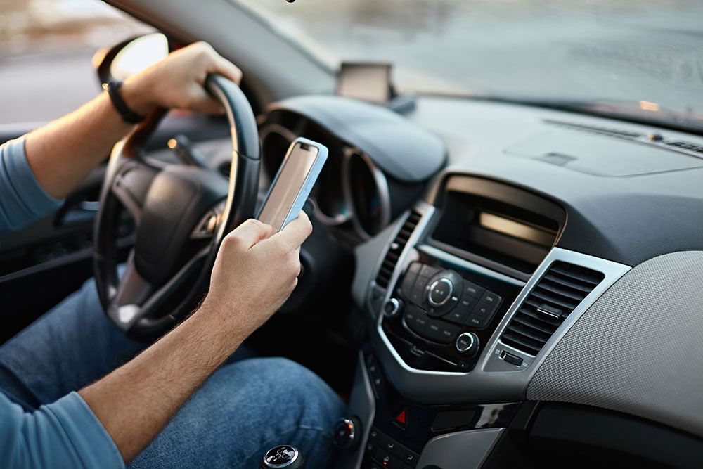 Driver holding a smartphone while steering a car, illustrating distracted driving hazards.
