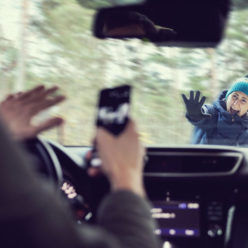 Distracted driver using phone, startled woman outside windshield, winter forest setting.