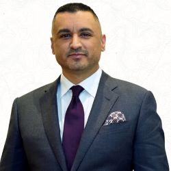 Business professional in a suit and tie, standing against a white background with a subtle pattern.