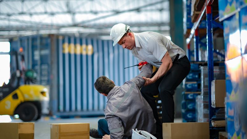 Warehouse safety: worker assists another after a fall among boxes and forklift. Safety helmets nearby.