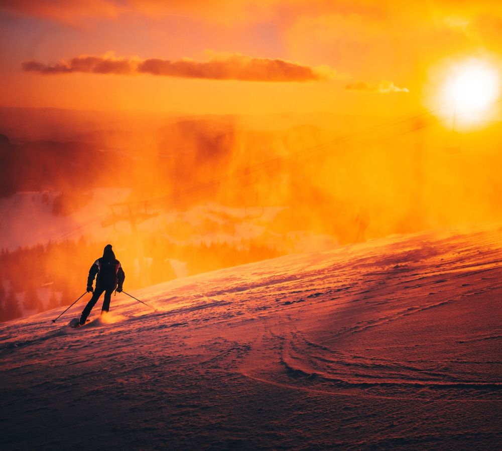 Skier descends snowy mountain at sunset, vibrant orange glow illuminating the scene.